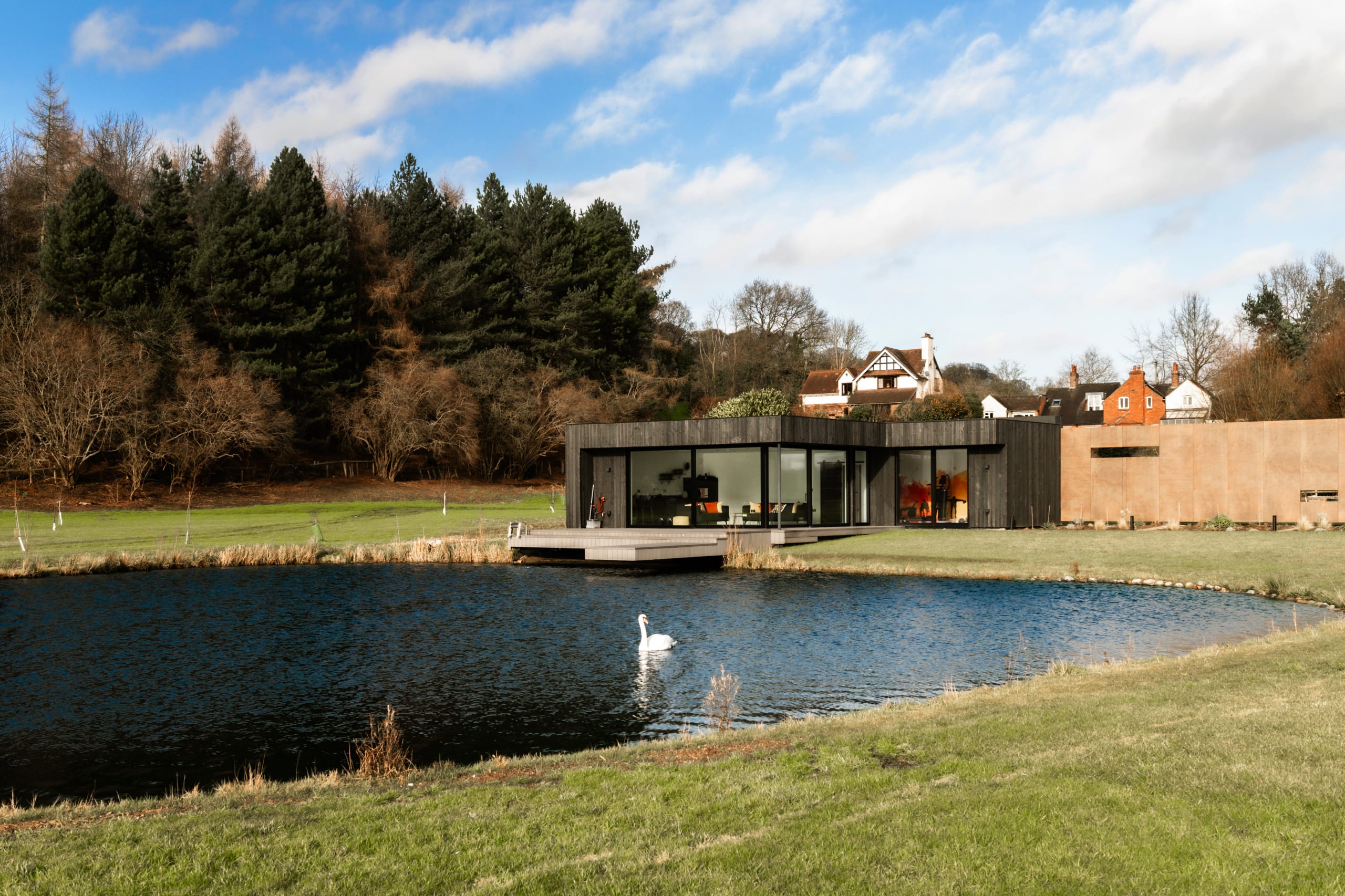 Modern single-story black timber-clad home with floor-to-ceiling windows overlooking a pond with swan, rural English setting